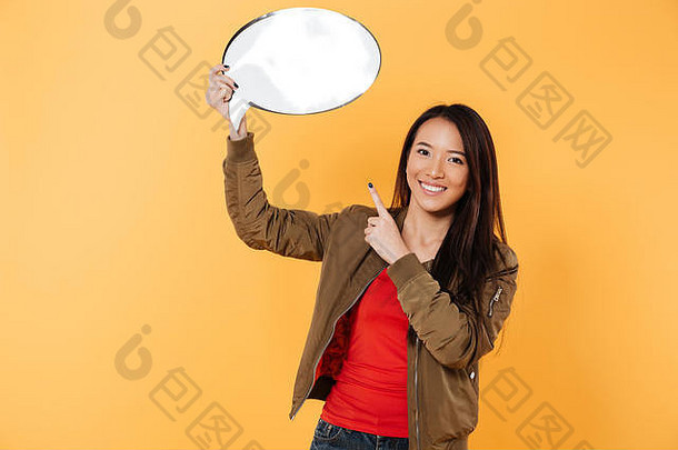 一个年轻的微笑的亚洲女孩的肖像，拿着空白的演讲泡泡，手指在黄色背景上孤立地指着
