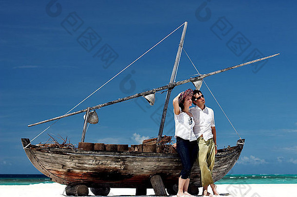在热带白沙海滩，一对幸福的年轻夫妇在老船旁摆姿势