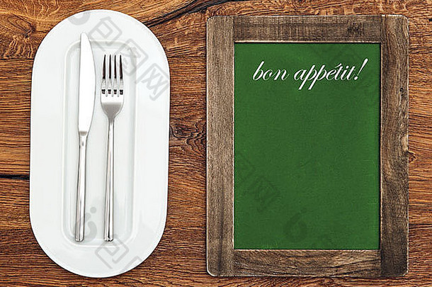 木桌上的绿色黑板和白色盘子、刀叉。示例文本bon appétit！