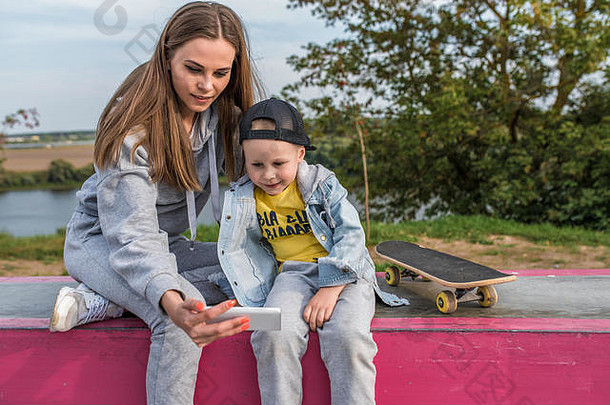 年轻的母亲和4-5岁的小男孩儿子，在夏秋时节在城市公园网站上，在手机上看视频，在互联网上在线申请