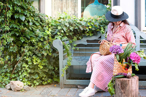 一个穿着衣服的漂亮女人，坐在城市街道的长椅上。一个女孩手里拿着一大篮蔬菜和花。空间。横幅