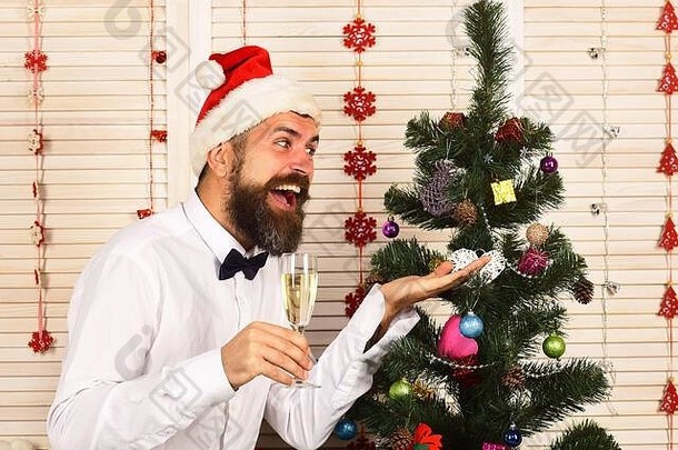 留着胡子的男<strong>人</strong>拿着香槟酒杯，指着枞树。庆祝和新年概念。木墙背景上圣诞树附近的家伙。戴着红帽子、脸上洋溢着欢乐的圣诞老<strong>人</strong>发出<strong>欢呼声</strong>