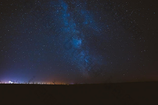 暗夜星空背景。田野上方的蓝色星空和背景上的城市灯光。银河系的天然发光恒星。东区视野