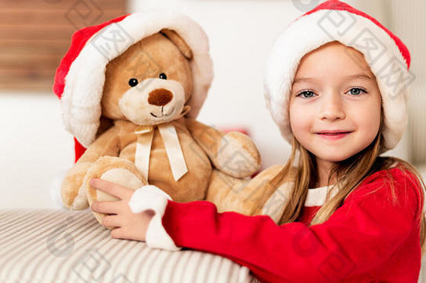 可爱的小女孩戴着圣诞帽，手里拿着她的圣诞礼物，软玩具泰迪熊。圣诞快乐的孩子，微笑着看着相机。