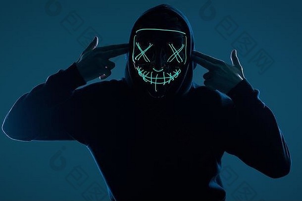 一个穿着黑色连帽衫的匿名男子的肖像，他的脸藏在可怕的霓虹灯面具后面。摄影棚拍摄。