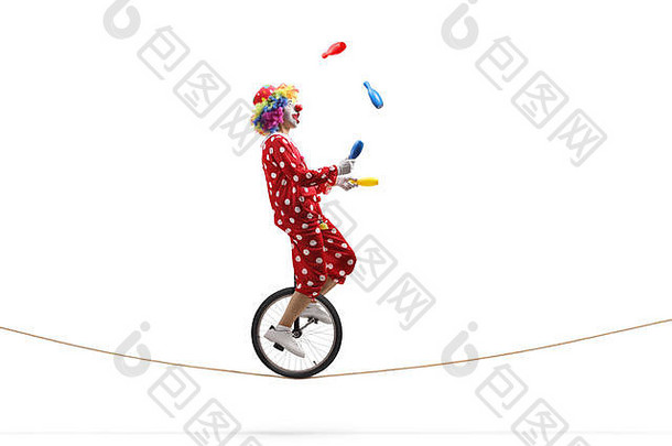一个小丑拿着棍棒玩杂耍，骑着一辆独轮车在白色背景上孤立的绳子上的全长侧面照片