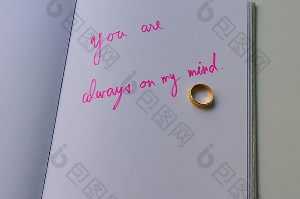 金色的戒指放在笔记本上，用粉红色的笔迹读着你永远在我的脑海里，书就放在白色的木桌上，爱的概念