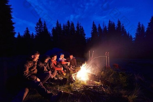 一群年轻人围坐在火炉旁，背景是大片云杉树和深蓝色的夜空。在森林里露营。度假与旅游理念