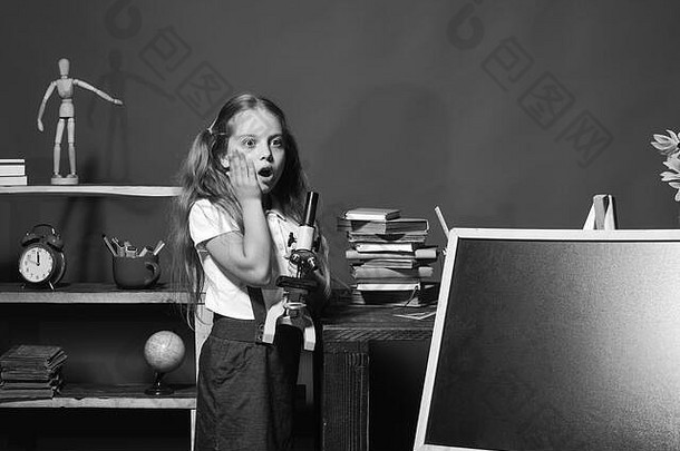 在教室里脸上带着震惊表情的女学生。女孩拿着显微镜，站在黑板旁，空间。教育问题与返校观念。红墙背景上的儿童和学校用品。