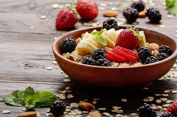 木制厨房餐桌上的粘土盘中有香蕉、草莓、杏仁和黑莓的水果健康慕斯里