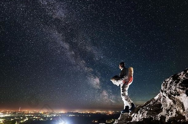宇航员站在岩石山上，美丽的夜空下有星星和银河。穿着宇航服的太空旅行者手持头盔，看着夜市灯光。宇航员和夜间的概念