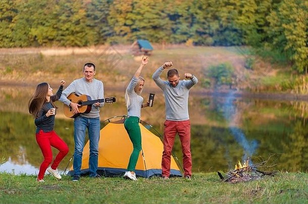一群快乐的朋友带着吉他，在户外玩得很开心，在公园的湖边跳舞和跳跃，背景是美丽的天空。露营的乐趣。