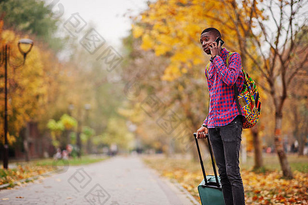 非洲美国男人。网纹衬衫手提箱背包黑色的男人。旅行者秋天公园说话移动电话