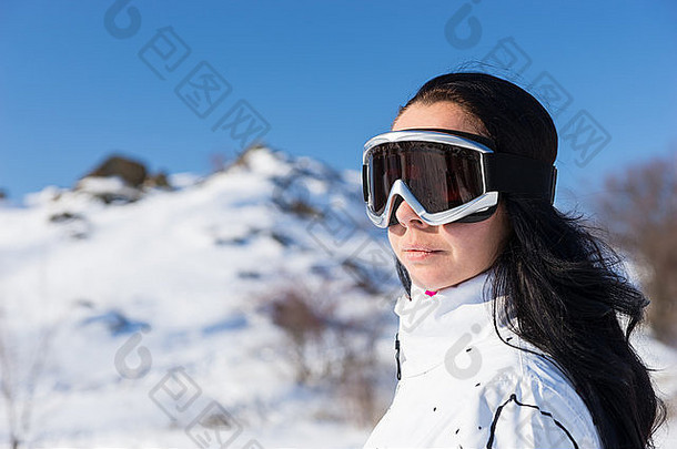 在阳光明媚的雪山上滑雪休息时，戴着滑雪镜、留着黑色长发的年轻女子的头像和肩膀特写，欣赏风景。