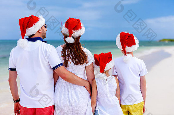 热带海滩上戴着红色圣诞老人帽庆祝圣诞节的美丽家庭的后视图