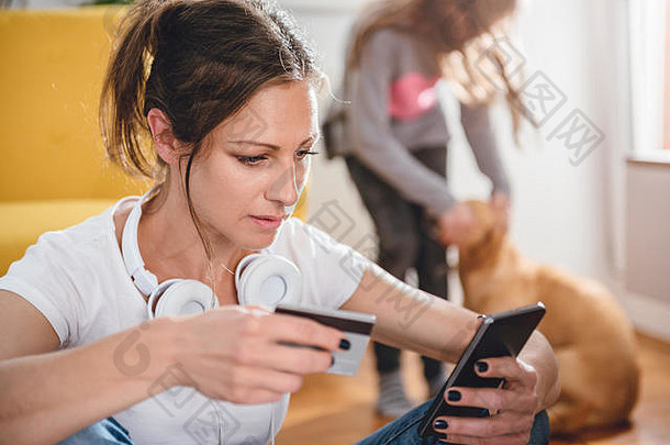 一名身穿白衬衫、戴着耳机的妇女在智能手机上网上购物，并使用，背景是一个女儿和一条狗