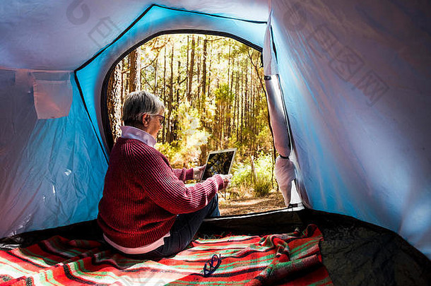 一位成年退休妇女坐在帐篷里，独自一人在森林里自由野营。她使用一种连接互联网的平板电脑来组织这次活动