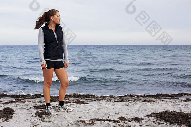 美丽的年轻女子站在海滩上眺望远方的全长图像。健康的女运动员<strong>跑步</strong>后放松