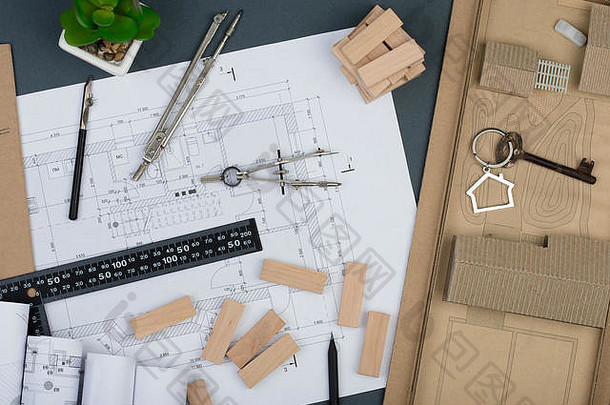 建筑师的工作场所-钥匙上有房子形状的小饰物、木块、建筑图纸、灰色背景上的工程工具