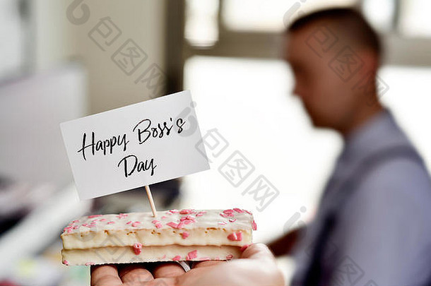 桌子上放着一块蛋糕的特写，上面有一块写着“老板日快乐”的招牌，上面有一个人在电脑里工作