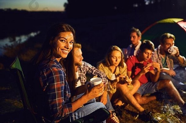 晚上，在篝火旁的朋友们的背景下，女孩端着一杯茶