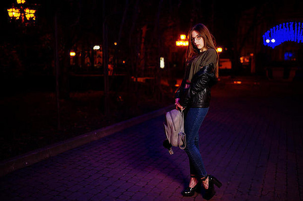 女模特戴眼镜、牛仔裤和皮夹克，手拿背包，在城市街道的蓝色灯光花环衬托下的夜照。