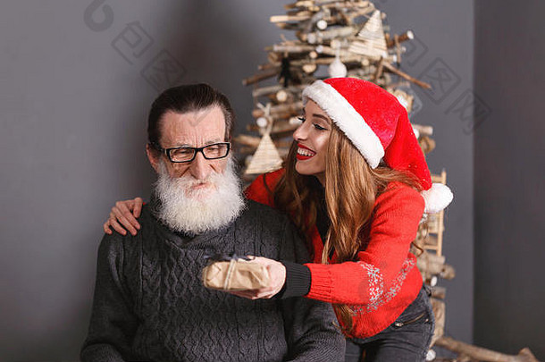 英俊的大胡子、戴眼镜、穿灰色毛衣的老爸爸接受了他年轻的长发女儿的礼物，女儿穿着红色毛衣、戴着圣诞帽，他感到震惊和惊讶，新年、圣诞节、假期、纪念品、礼物、<strong>购</strong>物、折扣、商店、雪人圣诞老人、化妆、发型、<strong>狂欢</strong>