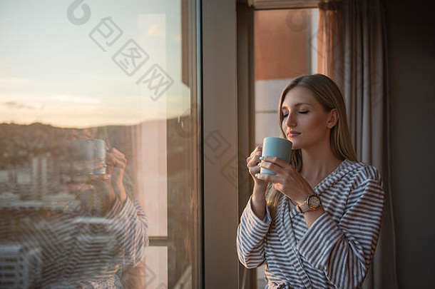 年轻的女人浴袍享受晚上咖啡美丽的日落景观城市站窗口