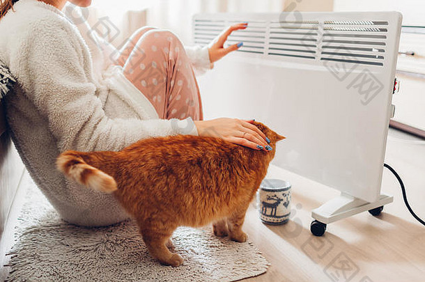 冬天在家使用取暖器。一名妇女穿着暖和的衣服，坐在装置旁用猫暖手。取暖季节。