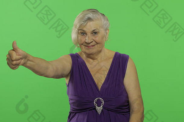 一位老妇人竖起大拇指微笑着。穿着紫色连衣裙的漂亮老奶奶。放置您的徽标或文字。色度键。绿色屏幕背景