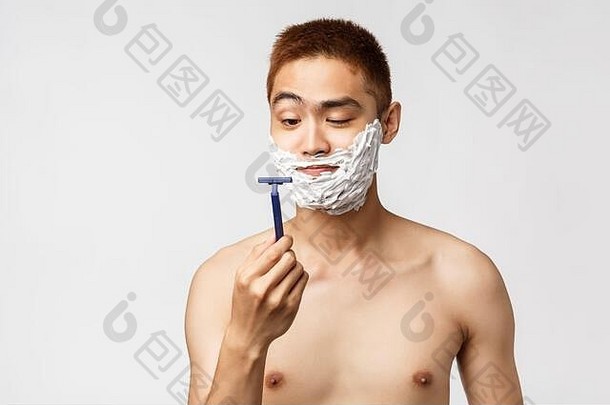 美、人、卫生理念。好奇英俊的亚洲男人看着新剃须刀开始刮胡子，脸上涂着奶油，光着身子站着
