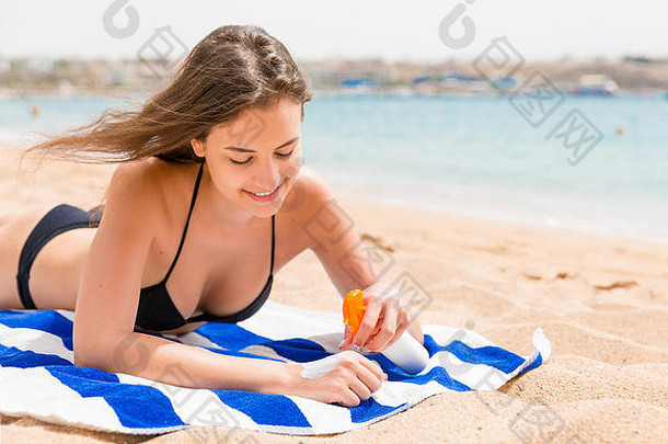 年轻快乐的女人在海边的毛巾上休息，用<strong>防晒</strong>霜保护手上的皮肤不受<strong>喷雾</strong>的伤害。
