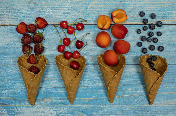 浆果和水果冰淇淋。在蓝色木质背景上，将各种新鲜水果、蓝莓、草莓、樱桃、杏平放在华夫格蛋卷中。夏季甜点菜单概念。冰淇淋制作
