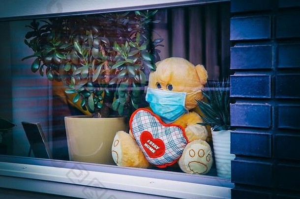 在家隔离，预防冠状病毒大流行。可爱的泰迪熊戴着医用面具，抱着心脏，坐在窗台上，旁边是有壳的罐子