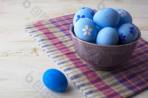 浅蓝色复活节彩蛋放在白色木质背景的格子餐巾上的紫色碗中。复活节背景。复活节彩蛋。东