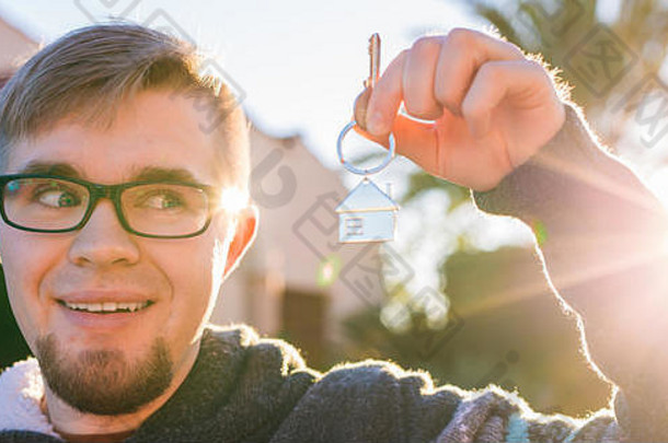 一个年轻有趣的男人拿着一个新家前面的房子形状的钥匙链上的房子钥匙。