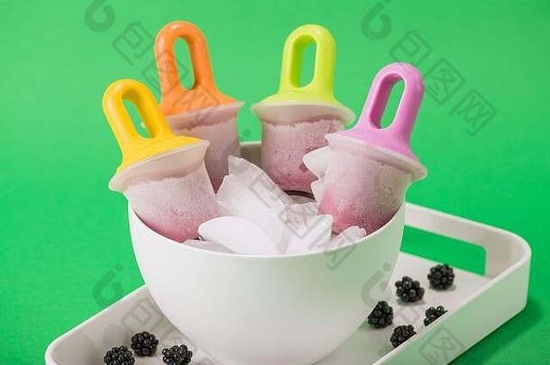 四个的黑莓冰棒放在一碗冰里，放在一个白色托盘里，托盘里放着新鲜的黑莓，背景是绿色的