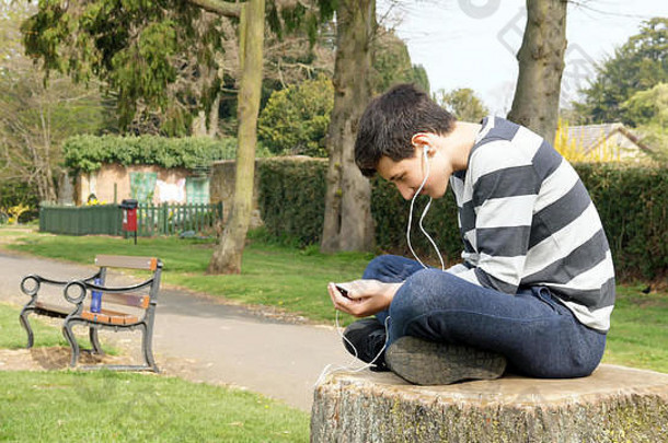 坐在英国公园的树桩上用智能手机听或说的十几岁男孩。青少年和社交媒体概念。