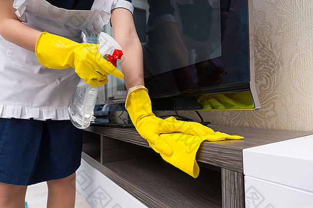 清洁电视柜的女佣在用布擦拭之前先喷上洗涤剂，然后用戴着手套的手靠近