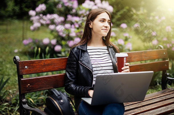 放松年轻的女孩研究春天公园坐着木板凳上咖啡浏览移动PC