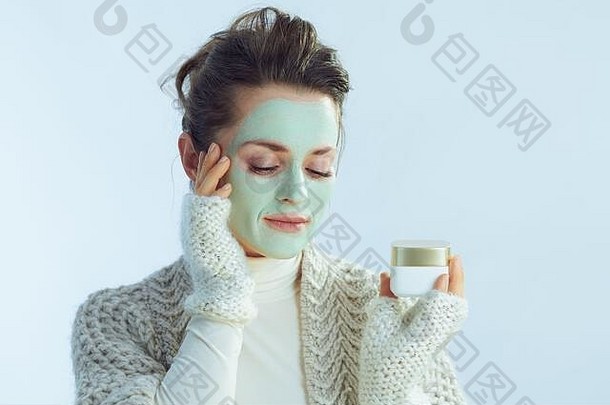 时尚的年女人卷脖子毛衣开襟羊毛衫绿色面部面具显示化妆品Jar冬天光蓝色的背景