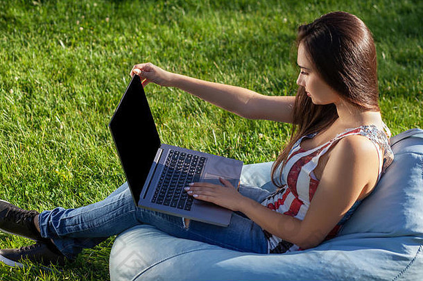 年轻的可爱的女孩移动PC坐豆袋花园公园绿色草在线购物概念