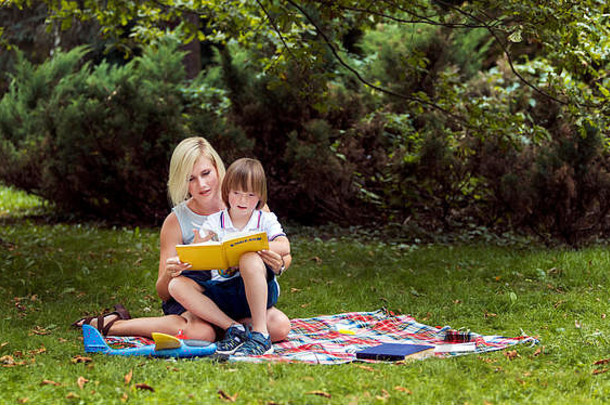 年轻的妈妈。阅读书特殊的儿子地毯在公园草坪上