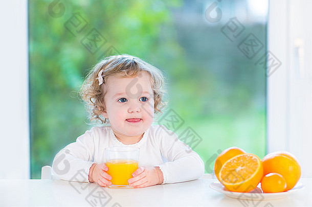 快乐微笑蹒跚学步的女孩喝橙色汁坐着白色表格大窗口