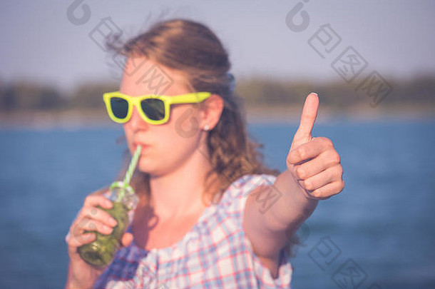 年轻的现代女人太阳镜喝<strong>猕猴桃</strong>汁显示拇指海边夏季概念