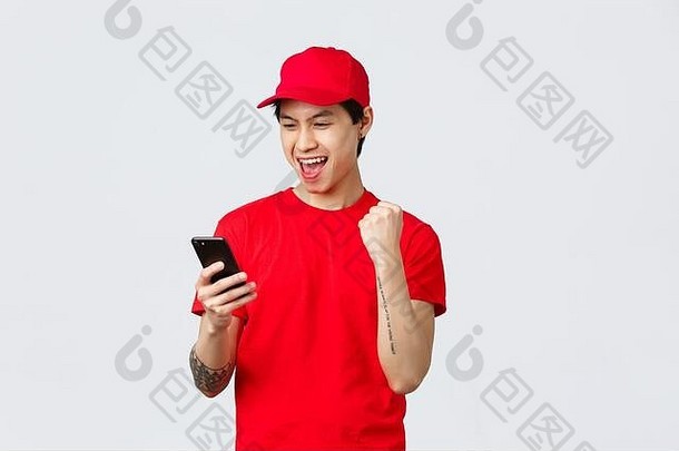 快乐年轻的亚洲交付的家伙红色的统一的t恤帽阅读好新闻智能手机屏幕使拳头泵高喊伟大的奖金成功