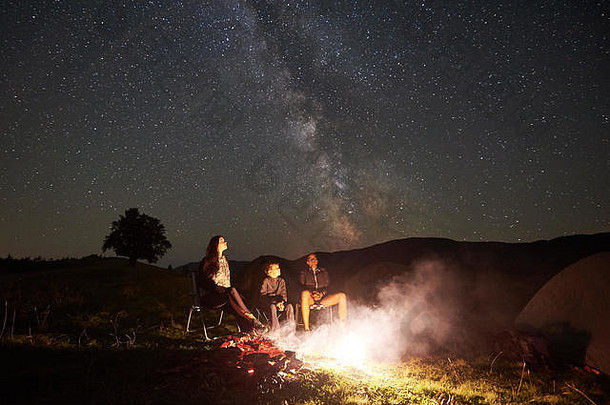 年轻的旅游家庭休息山坐着椅子营篝火帐篷夏天晚上妈妈。父亲儿子享受晚上布满星星的天空完整的星星乳白色的