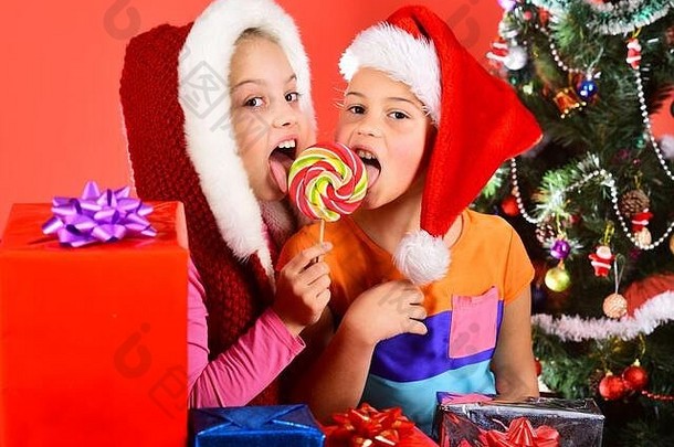 女孩庆祝圣诞节姐妹圣诞节帽子礼物盒子开放礼物聚会假期概念孩子们很高兴脸吃糖果红色的背景