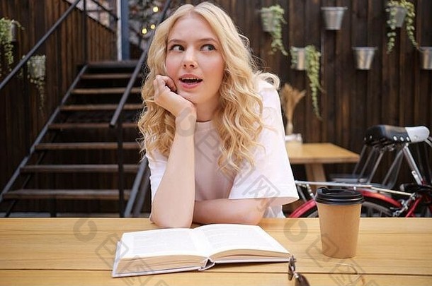 有吸引力的使惊讶金发碧眼的女孩倾斜手惊奇地书咖啡院子里咖啡馆