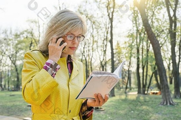 户外肖像成熟的业务女人眼镜阅读书会说话的电话女金发女郎黄色的雨衣背景春天公园